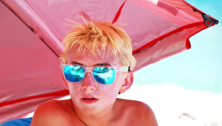 teen wears sunscreen and mirrored sunglasses under red beach umbrella