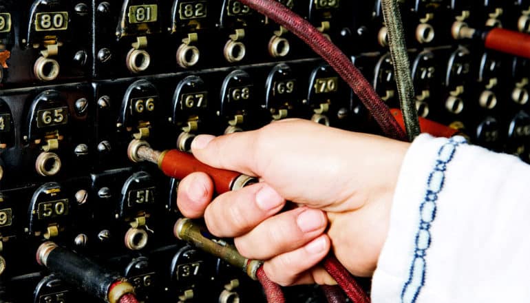 A person at an old telephone switchboard pulls one red plug out of the board