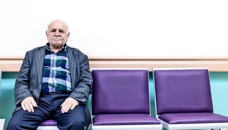 An older man sits on a bank of purple chairs in a waiting room