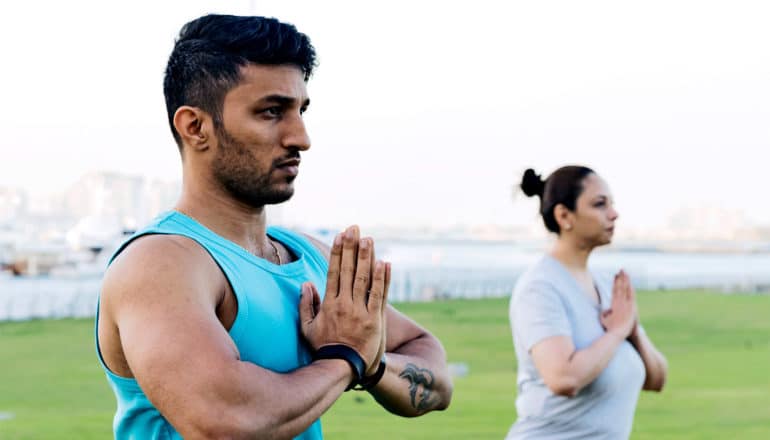 A man in a blue tank top holds his hands together while doing yoga outside, with a woman in the same pose in the background