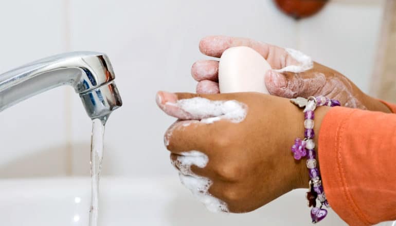 A child washes their hands in a white sink with a bar of soap. They have on a charm bracelet and an orange shirt