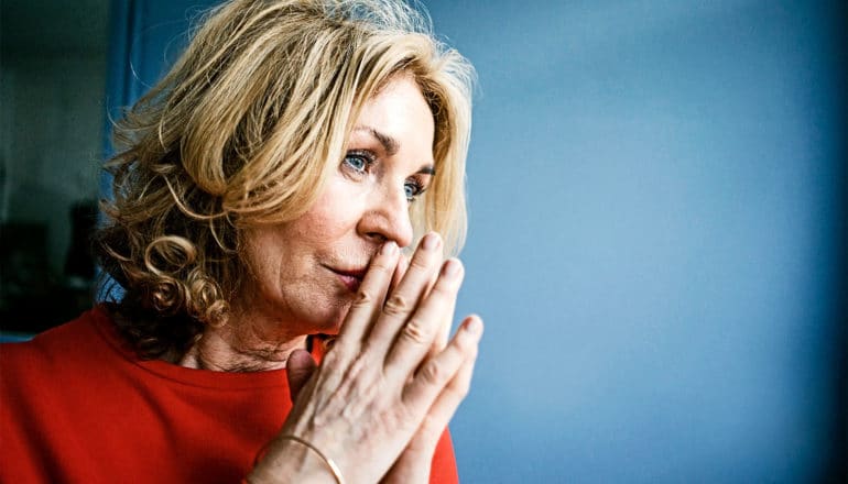 An older woman in red looks serious with her hands to her mouth, lost in thought, with a blue background behind her