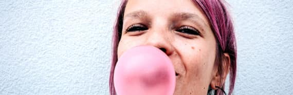 A young woman with pink hair blows a pink bubble with chewing gum while standing against a white wall