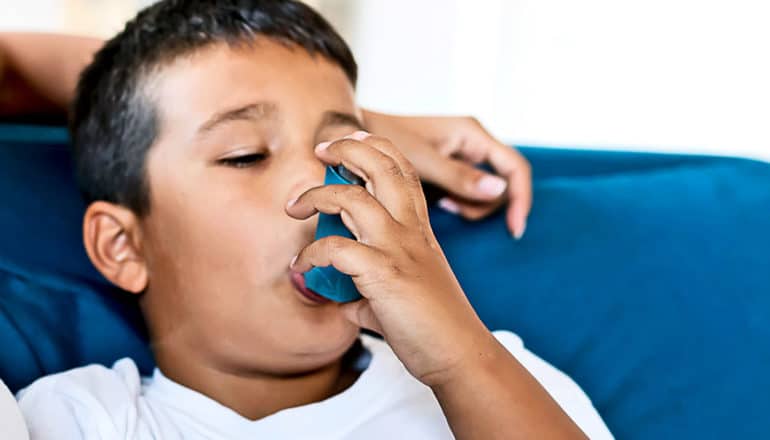 A young boy uses an inhaler as he sits on a blue couch