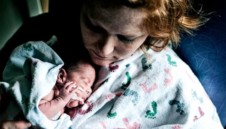 A mother holds her premature baby under a blanket covered in baby footprints. The baby has tubes coming out of its nose