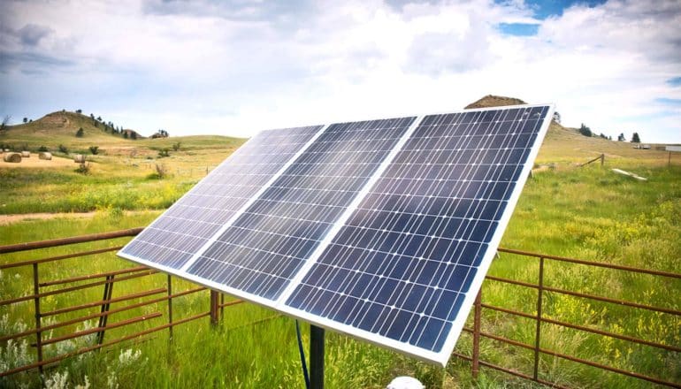 A solar panel sits in a field with mountains in the background and a cloudy sky above