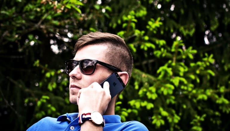 A man in a blue polo shirt and sunglasses talks on a cell phone against a background of green leaves