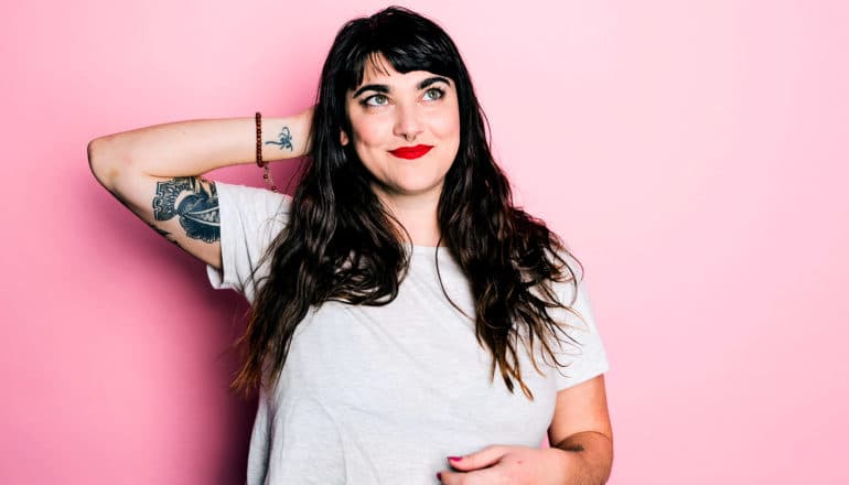 A young woman looks pleased with herself as she scratches the back of her head and looks up against a pink background