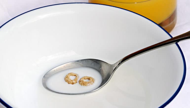 Two Cheerios touch in the milk inside a spoon sitting in an empty cereal bowl, with a glass of orange juice in the background