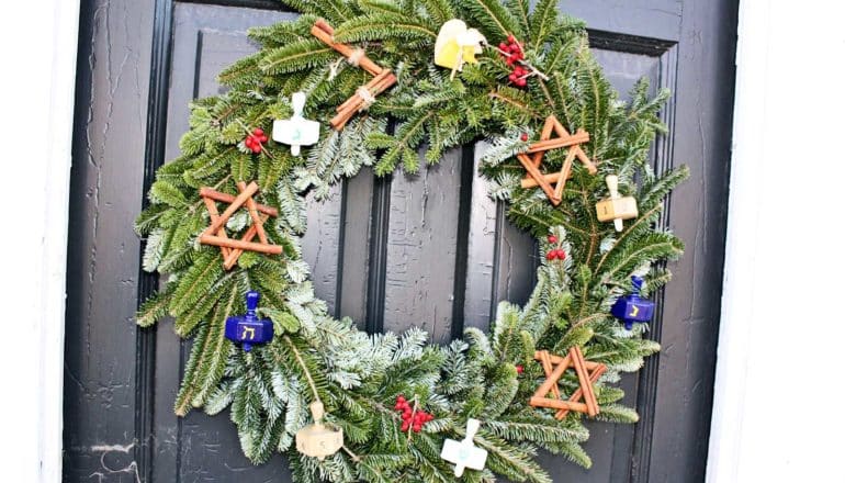 A wreath on a dark door has dreidels hanging in it alongside cinnamon sticks in the shape of the Star of David
