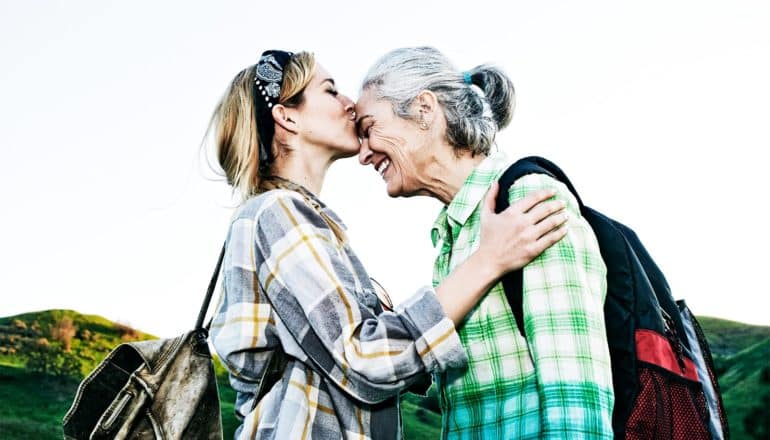 A young woman wearing a flannel shirt and a backpack kisses an older woman on the forehead as she smiles, with mountains and sky behind them