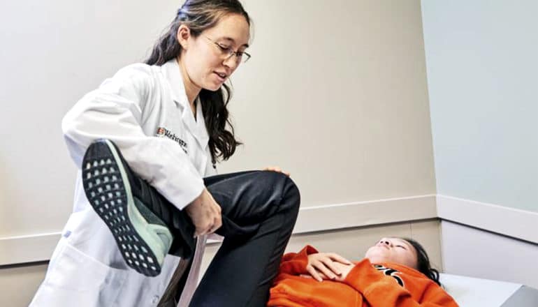 A doctor checks a young patient in an orange hoodie, lifting her leg up while she lays down