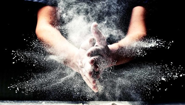 A person claps their flour-covered hands together, sending flour flying in every direction, all against a dark background