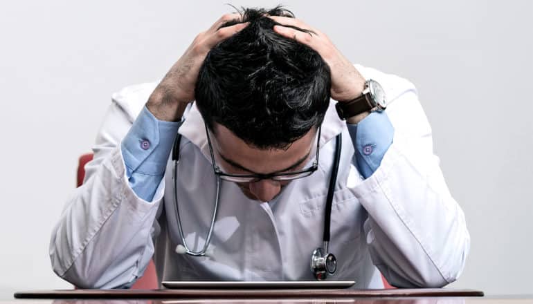 A young doctor in a blue shirt and white coat looks down at an iPad with his hands on the back of his head in exasperation.