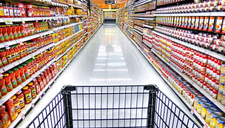 A shopping cart points down a grocery store aisle lined with canned and jarred foods