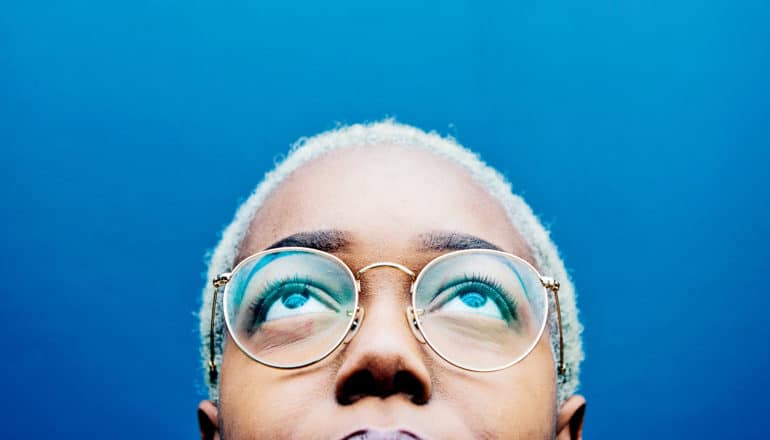 A young woman with glasses looks upward, with only her face above her mouth visible, against a blue background
