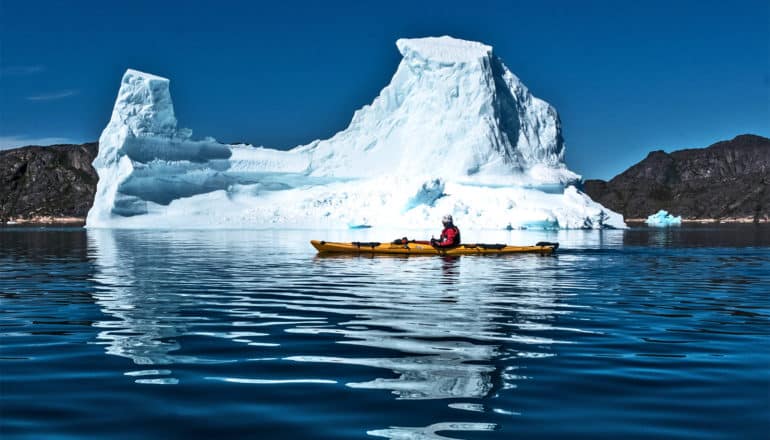 A kayaker moves past a large glacier on deep blue water, with rocky crags behind it