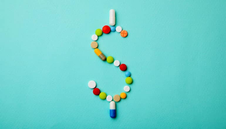 Pills of many different colors are arranged into a dollar sign sitting on a blue/green background