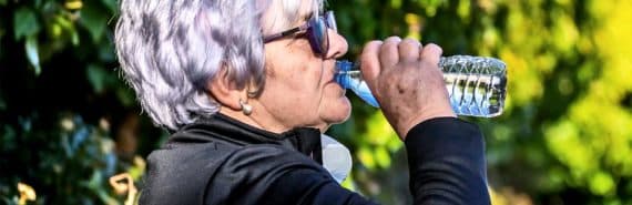 An older woman drinks from a water bottle while sitting on a park bench wearing a black top and sunglasses
