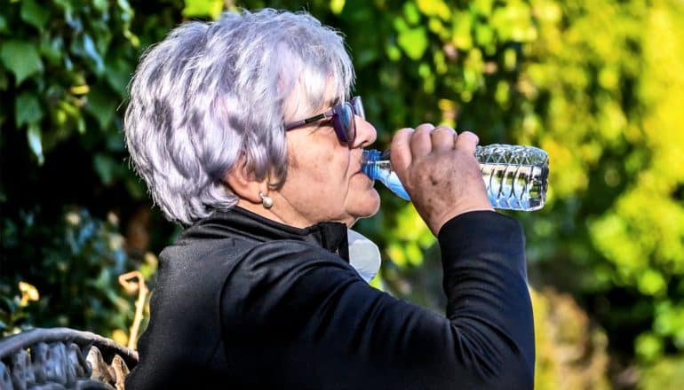 An older woman drinks from a water bottle while sitting on a park bench wearing a black top and sunglasses