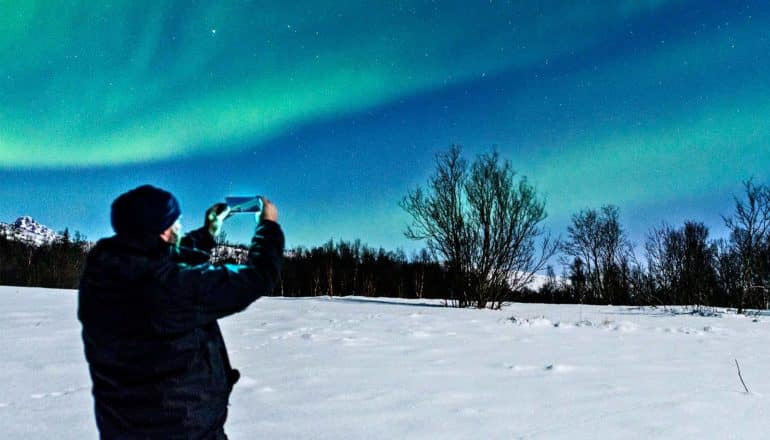 A man in a dark coat and hat stands on a snowy field using his phone to take a picture of the starry sky, filled with the green light of the Aurora Borealis