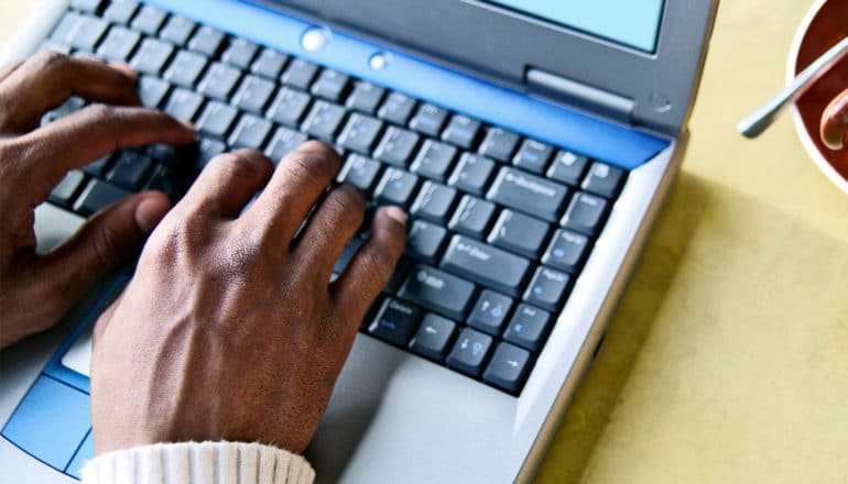 Hands sit on an old laptop computer keyboard on a yellow table