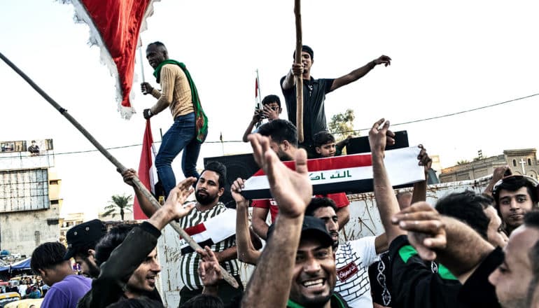 Protesters raise their hands and wave the Iraqi flag while standing above a crowd