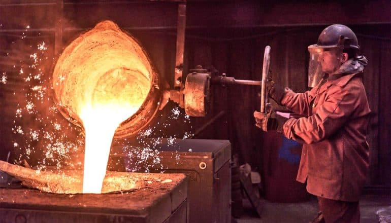 A man in protective gear turns a valve to pour a vat of liquid metal into another vat