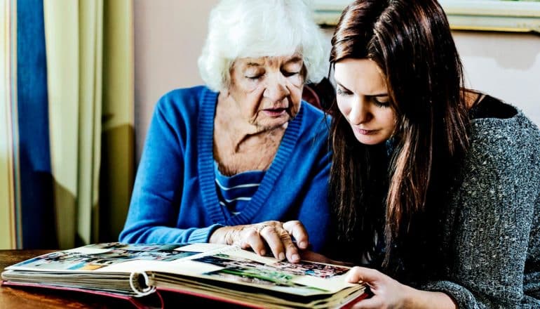 A grandmother in a blue sweater looks at a photo album with her grandaughter, who's wearing a dark gray sweater