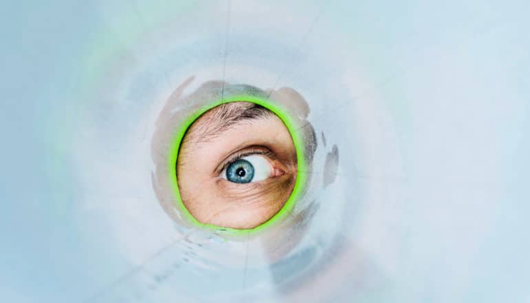 A person peeks through a tube with one eye which looks silvery on the inside and has a green ring around the opening