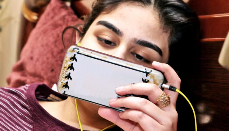 A teen wearing headphones watches a movie on her phone while laying down