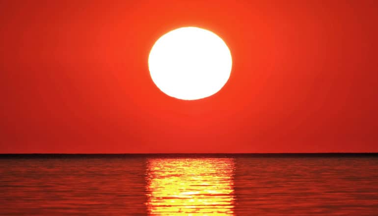 The sun sets over water, appearing totally white against a deep red sky with an orange reflection over water