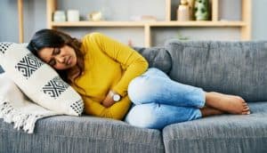 A woman in a yellow top and blue jeans lies on a couch with her hands on her stomach as she grimaces in pain