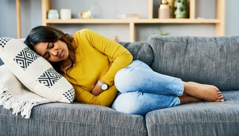 A woman in a yellow top and blue jeans lies on a couch with her hands on her stomach as she grimaces in pain