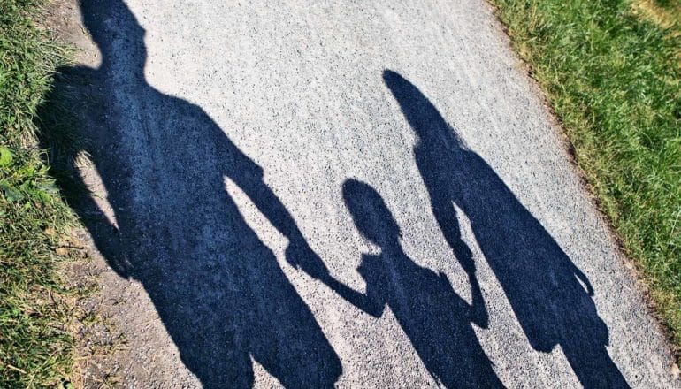 The shadows of parents hold their child's hand as they walk along a grass-lined path