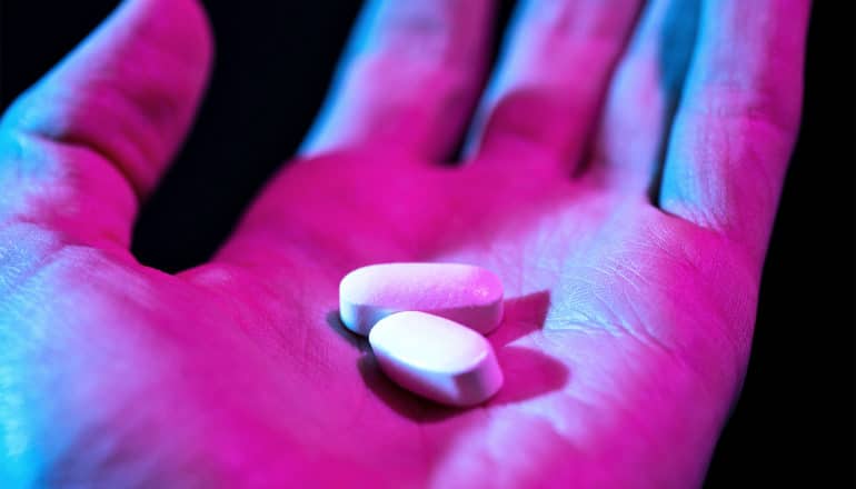 A person holds two white pills in the palm of their hand, covered in pink and blue light