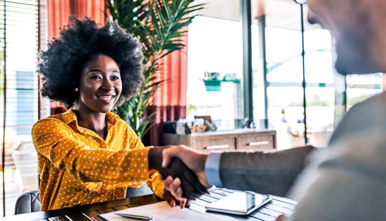 A young woman in a yellow shirt shakes the hand of a job interviewer inside a cluttered office with a plant behind her