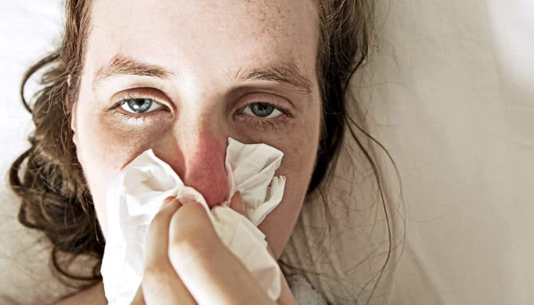 A woman with a cold blows her red nose while looking miserable on a white sheet