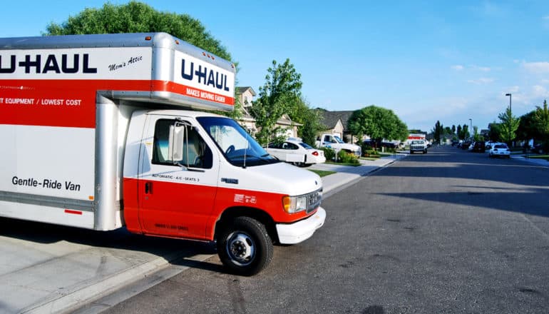 A moving truck is parked in a driveway on a suburban street with trees, other houses, and a blue sky in the background