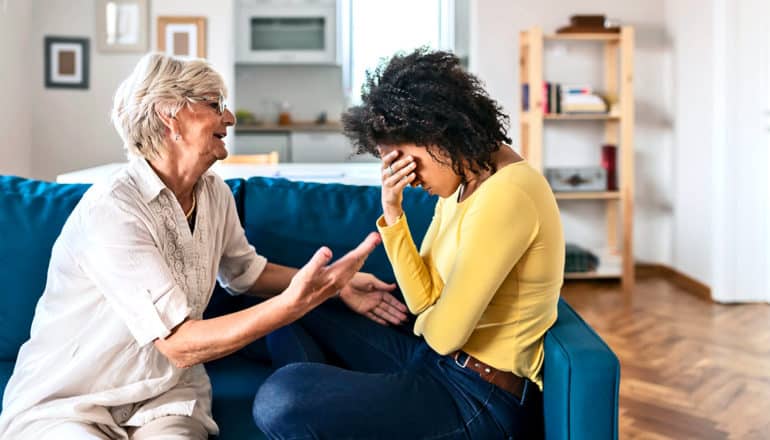 An older woman in white speaks to a younger woman in yellow who has her hand over her face as they sit on a blue couch in a white-walled apartment