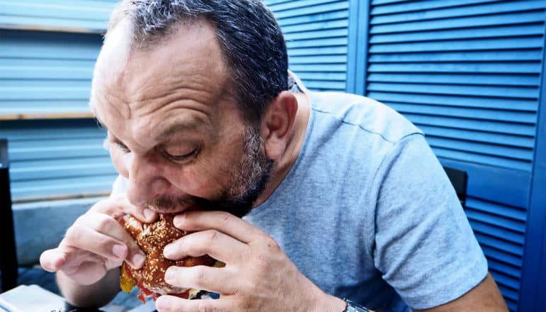 An older man in a gray t-shirt bends down over a table to take a big bite of a burger while sitting against a blue wooden window shade