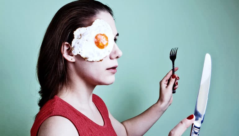 A young woman in red holds a fork and knife up while a fried egg hangs on her face