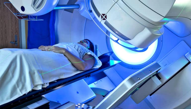 A woman in a hospital gown lays in a radiation therapy machine, bathed in blue light from the machine