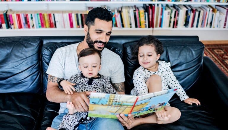 A dad in a gray t-shirt and blue jeans reads to his two young children on a black couch with a bookshelf in the background