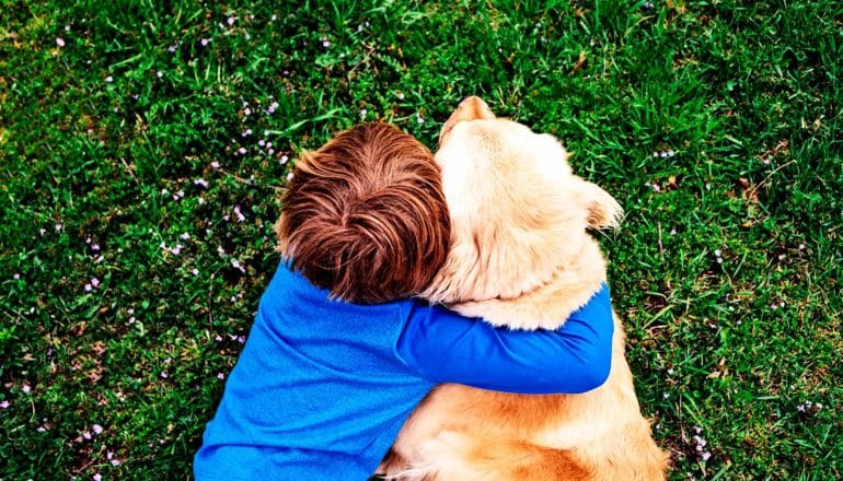 A red-haired boy in a blue shirt hugs a Golden Retriever as they lay on green grass