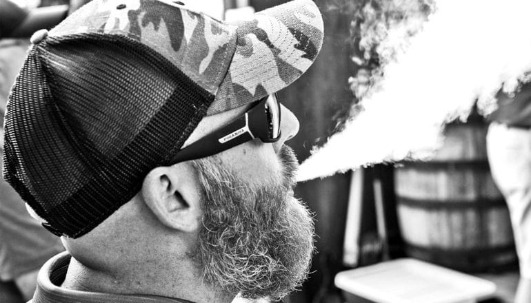 A bearded man in a hat and sunglasses blows out a cloud of vapor
