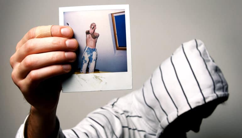 unrecognizable person in hoodie holds up polaroid photo of shirtless young man pointing gun at camera