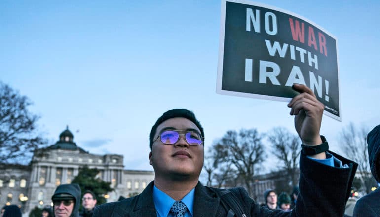A protestor in a suit and glasses holds a black and white sign that reads "No War with Iran" with "war" in red