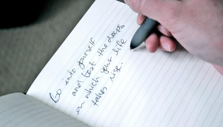 hand holds pen over notebook, where "Go into yourself and test the deeps in which your life takes rise" is written