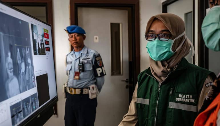 person in uniform and another in face mask and "health quarantine" vest in front of screen in airport